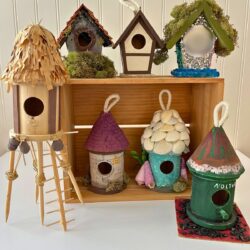 finished birdhouses, some stacked in a wooden box, some sitting on table, with bead board background.