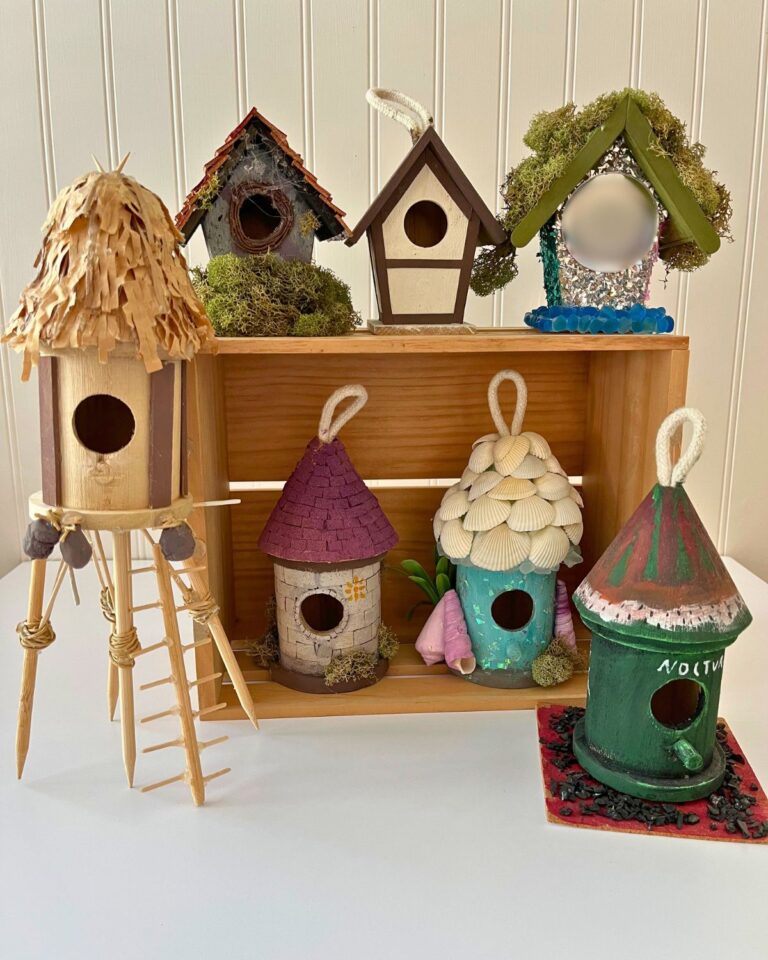 finished birdhouses, some stacked in a wooden box, some sitting on table, with bead board background.