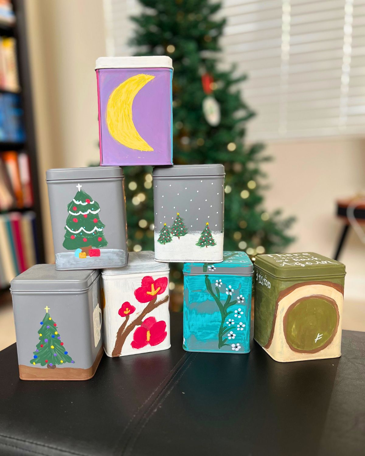 stack of painted tea tins on a black ottoman with christmas tree in the background.