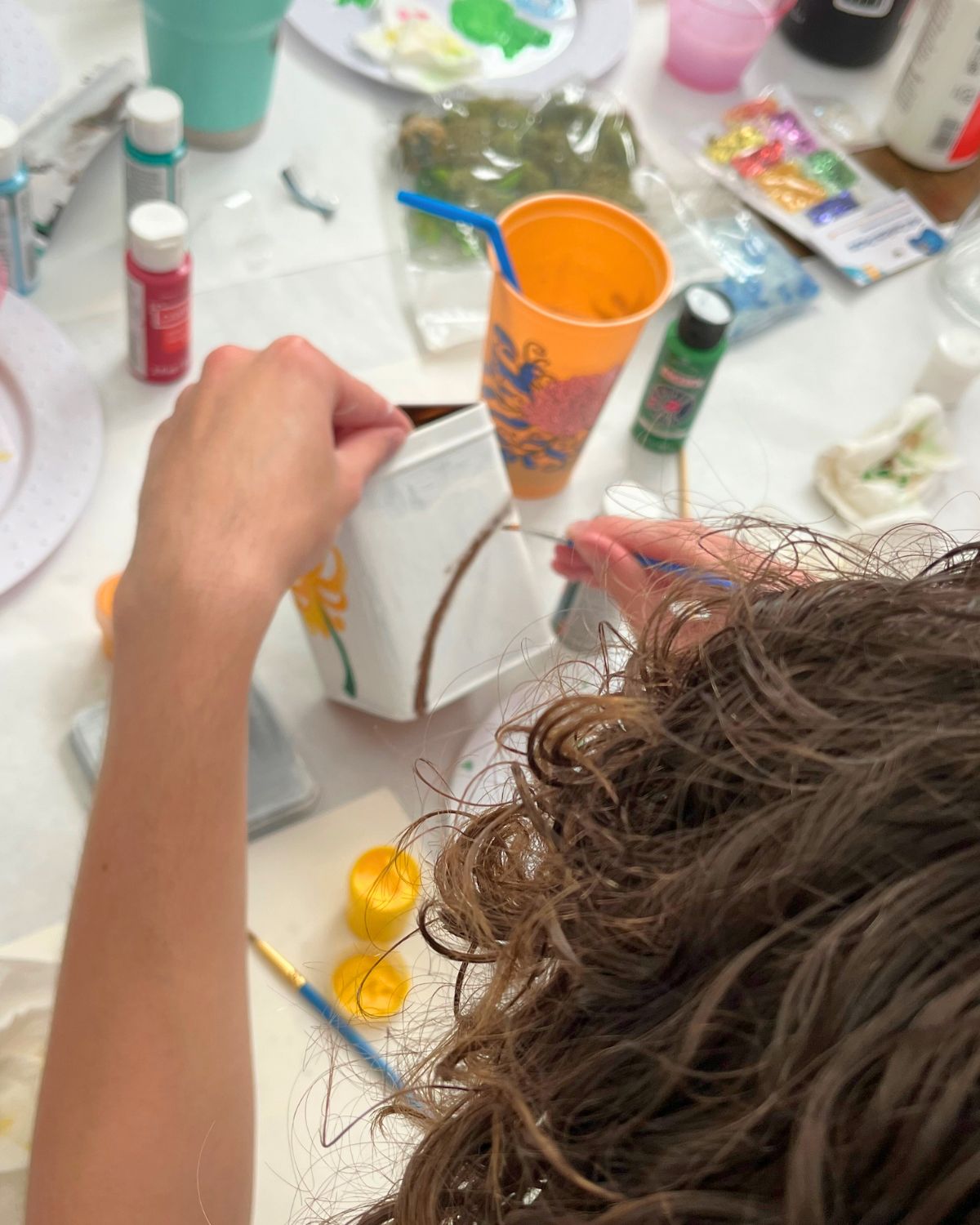view of a person painting a tea tin with supplies on table in front of him.