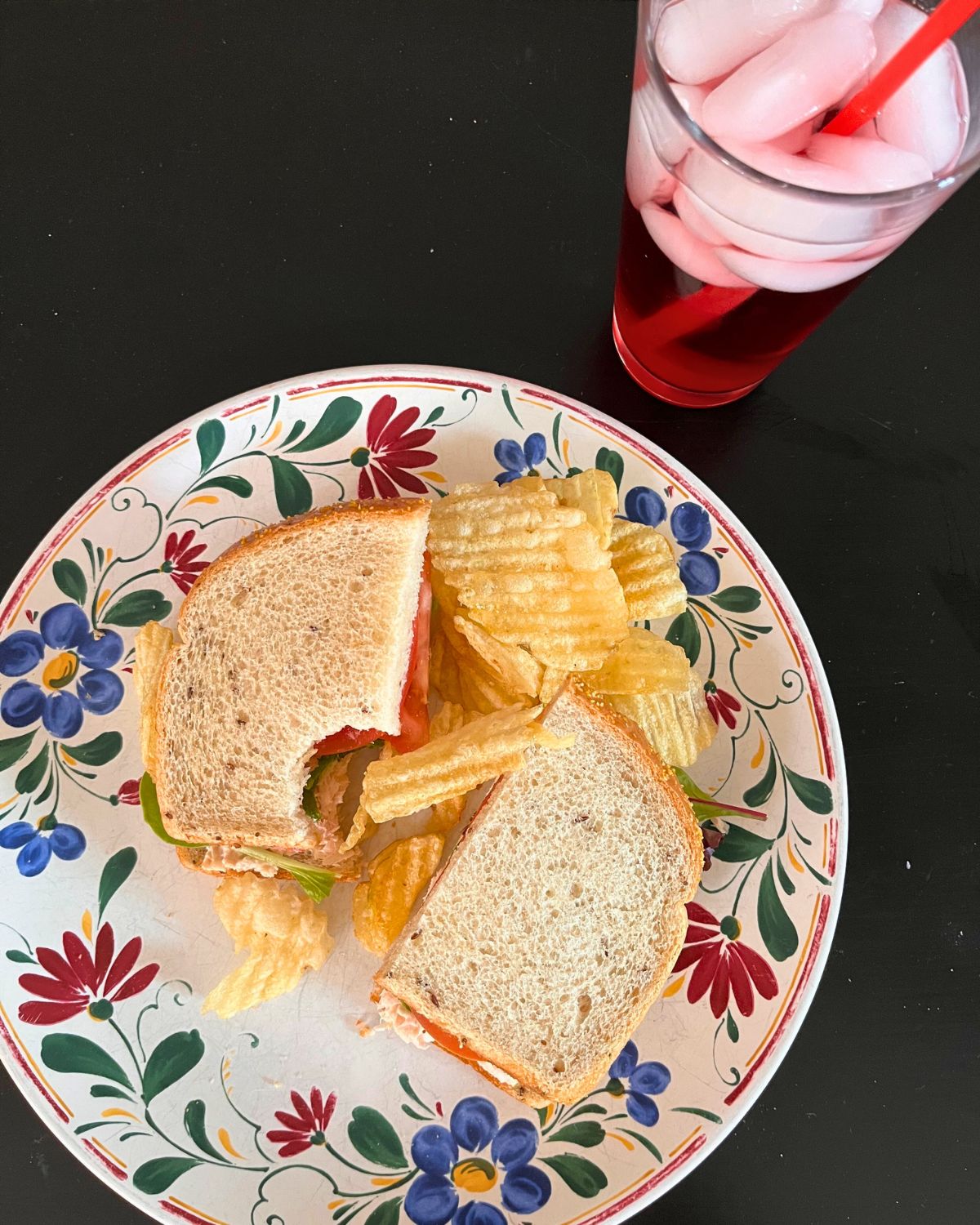 tuna sandwich on rye bread cut in half with a pile of chips on a plate with an iced red soda in a glass nearby.