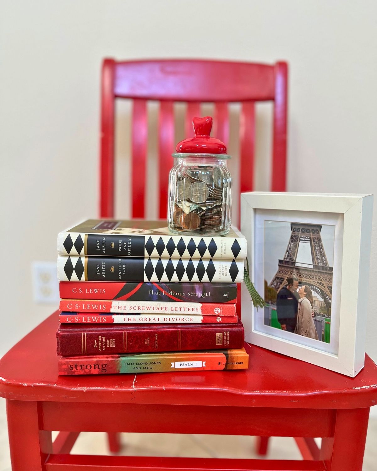 a red chair piled with books, picture frame, and jar of money in a heart container.
