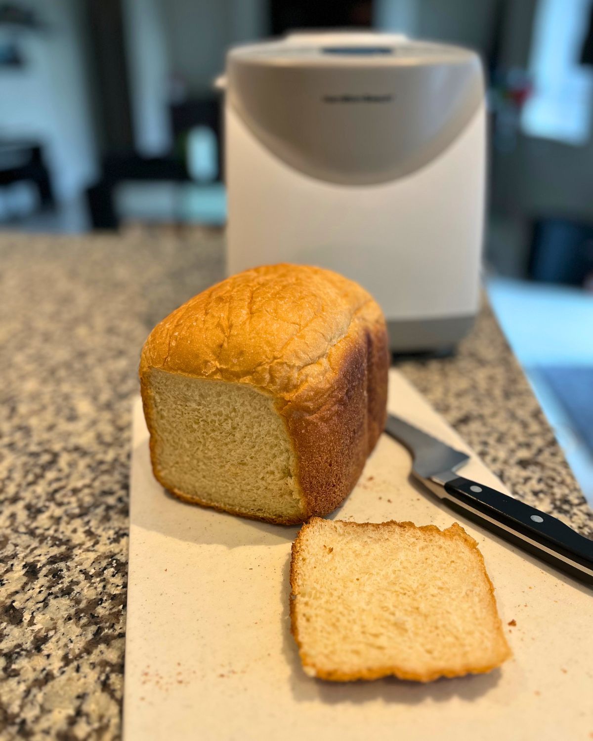 bread machine on counter with baked loaf on cutting board, sliced with a bread knife nearby.