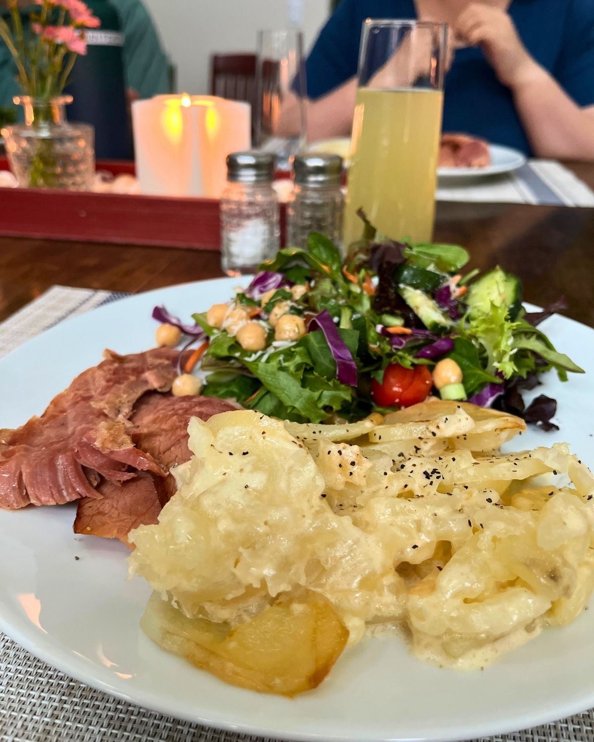 sunday dinner plate in foreground with champagne glass, salt and pepper, candles, and people in the background.