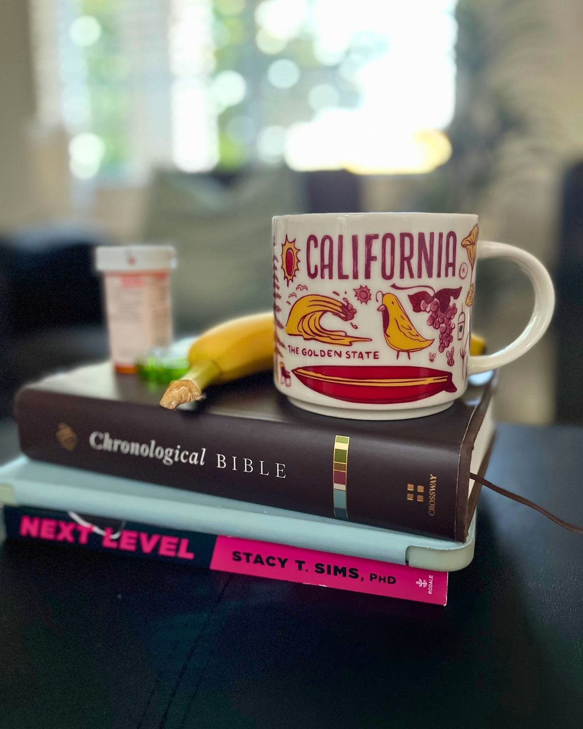 california mug atop bible, iPad, next level book, with a banana and medication.
