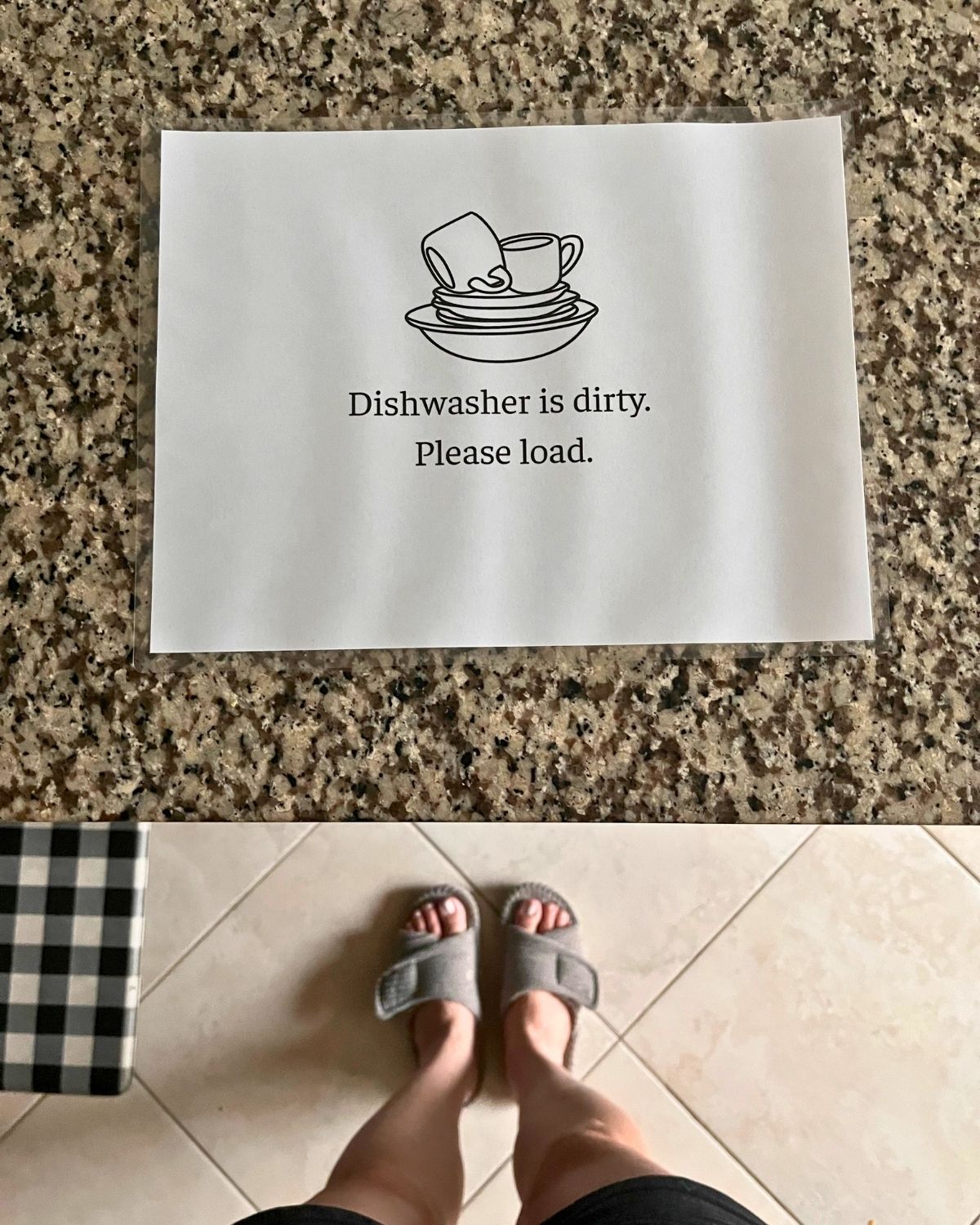 dirty dishwasher sign on counter with view of slippered feet and kitchen mat on floor below.