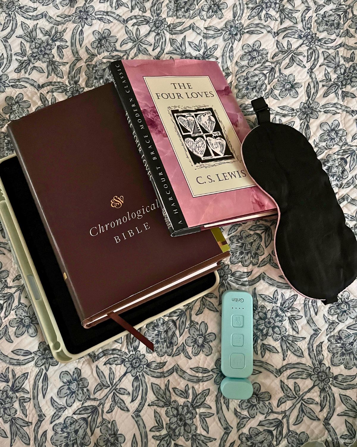 stack of books, iPad, mask, and reading light on bed.