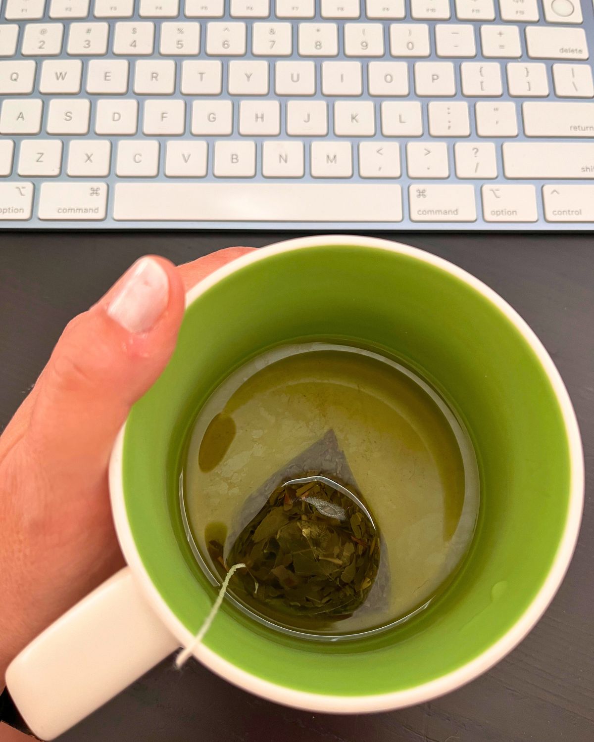 hand holding cup of tea by computer keyboard.